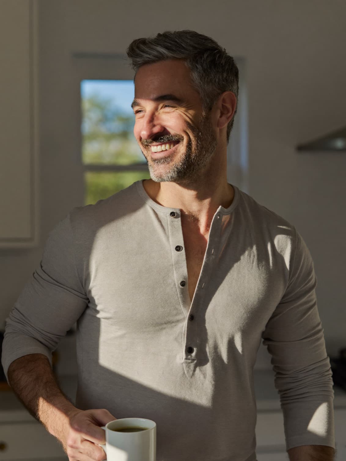 A man in his forties with salt-and-pepper stubble smiling in a sunlit kitchen