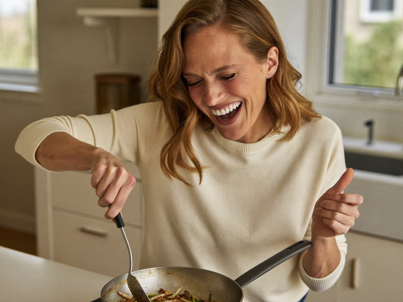 A woman in her forties laughing while cooking in a bright kitchen