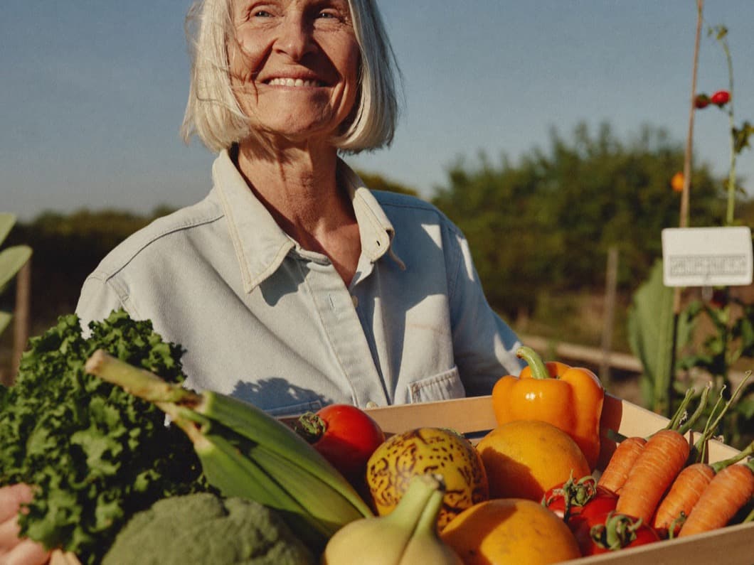 A woman in her sixties with a silver bob carrying a basket of vegetables in a garden