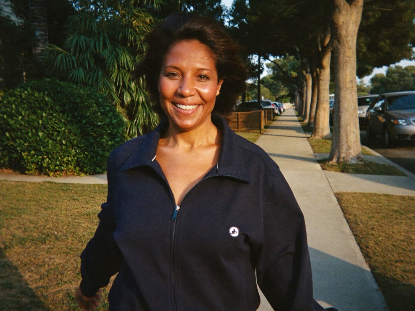 A woman in her fifties smiling as she walks down a tree-lined sidewalk in morning sun
