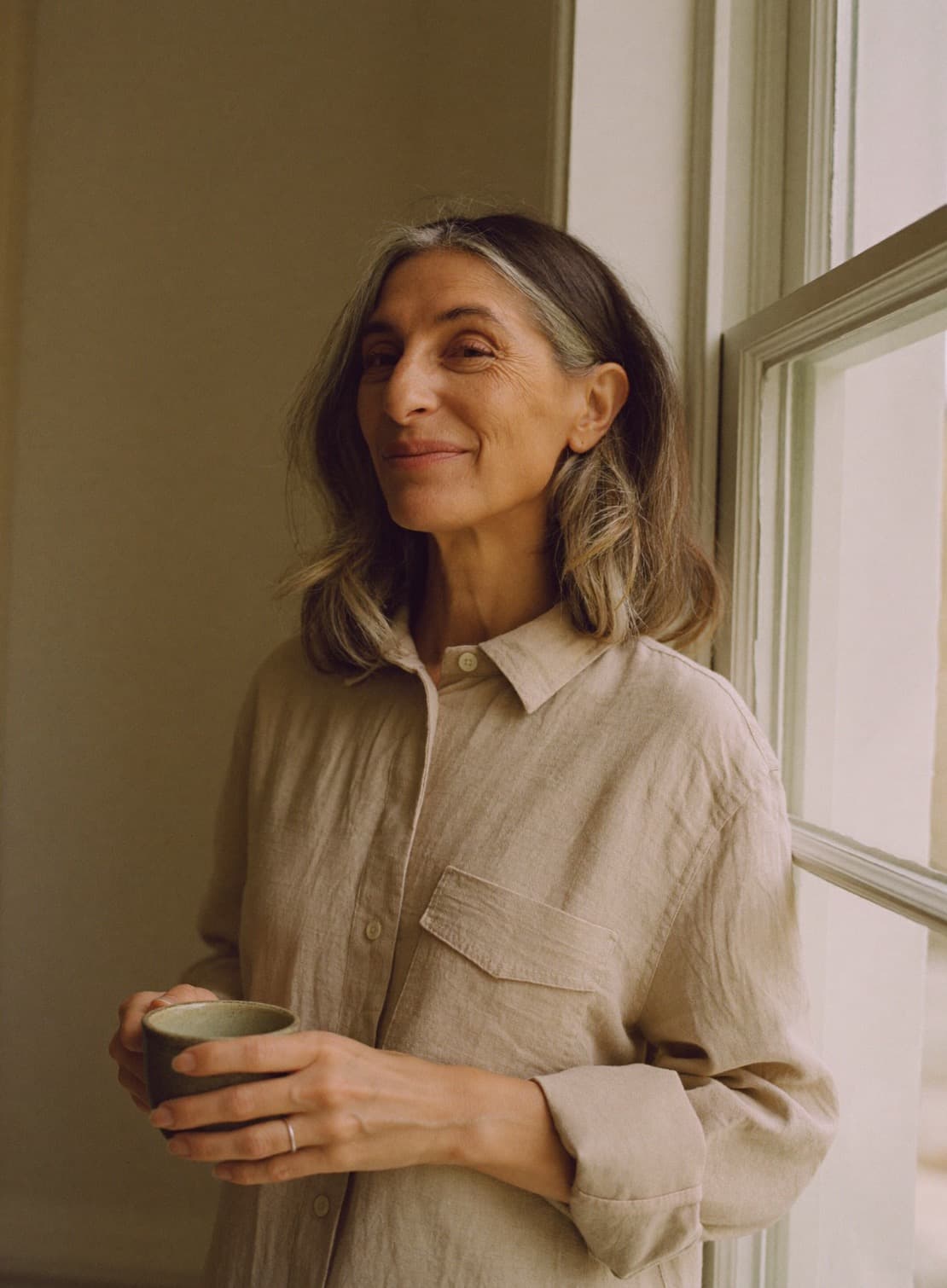 A woman in her late forties with silver-streaked hair smiling by a sunlit window