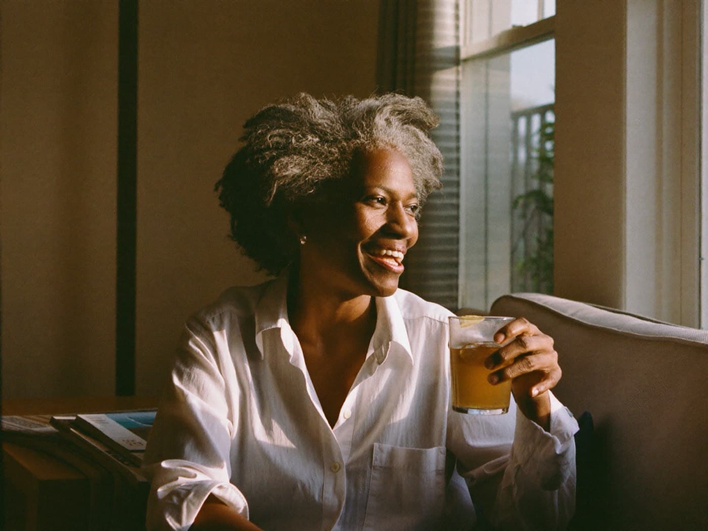 A woman in her fifties with gray natural hair laughing by a sunny window with a glass of iced tea