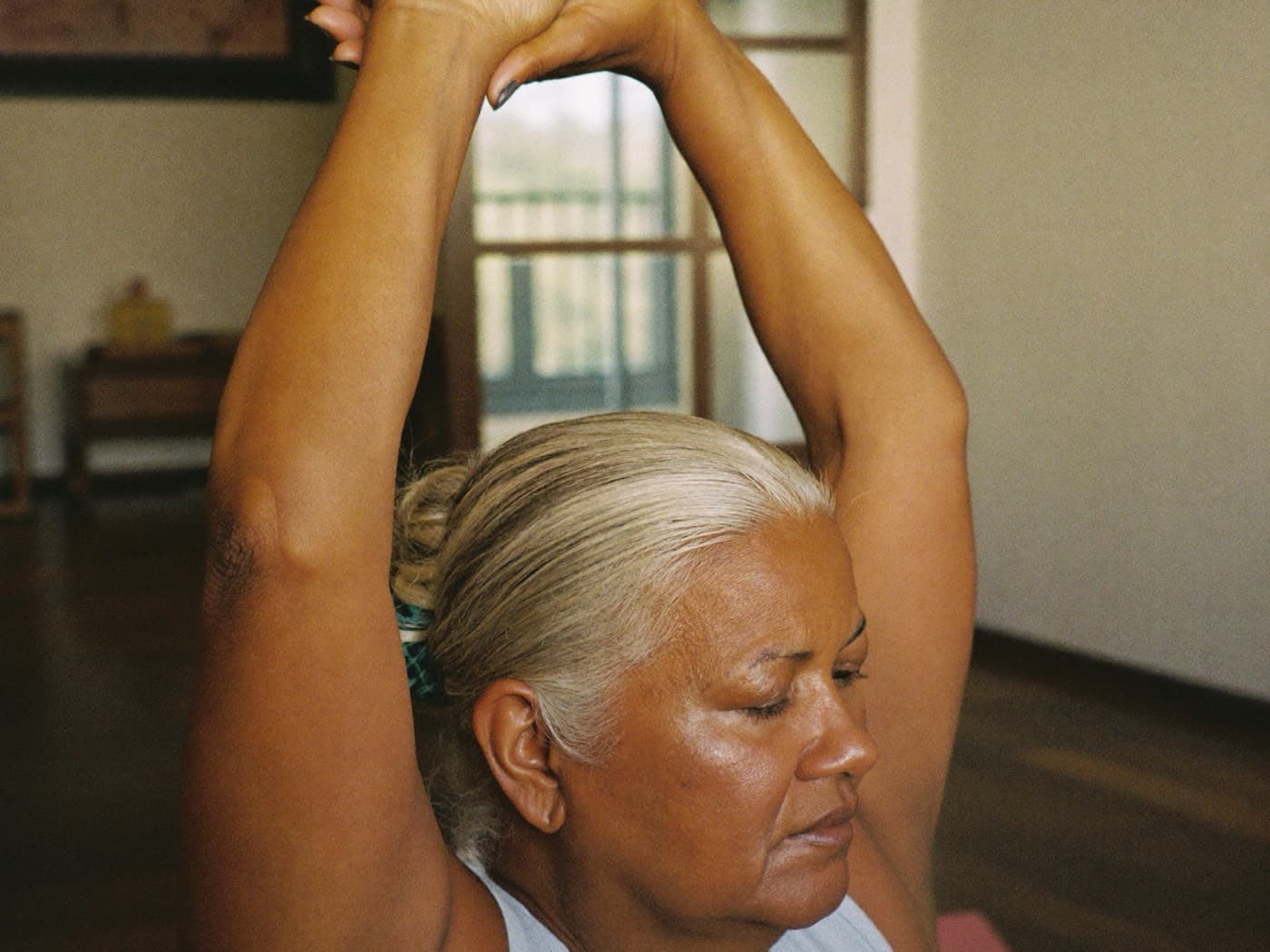 A woman in her sixties with silver hair in a low bun stretching her arms overhead at home